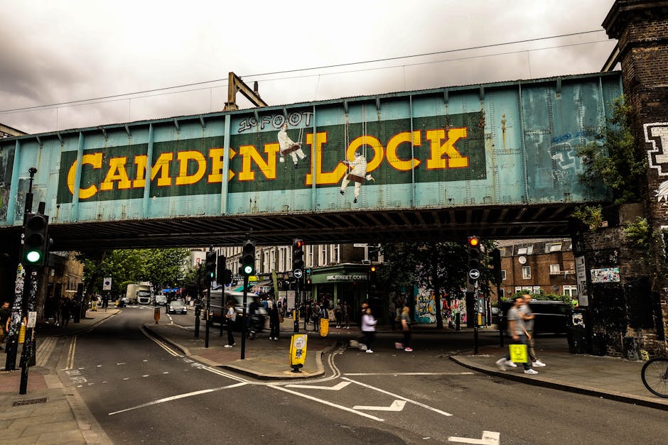 A large, weathered railway bridge spanning a busy street in Camden Town, Kentish Town, with the words 'CAMDEN LOCK' painted in bold yellow letters across its light blue-green metal surface. The bridge features graffiti art, including two clinging figures painted on the sides. Below, the street scene includes traffic lights, with one showing a green signal, and a variety of pedestrians walking along the pavement. Several small, portable traffic cones and signs are positioned on the road, indicating a possible controlled or restricted access area. In the background, a mix of brick buildings, shopfronts, and street-side murals can be seen, along with parked vehicles and moving traffic, creating a typical urban environment. The scene is illuminated by natural daylight under an overcast sky, capturing a moment of home relocation or furniture transport activity involving the surrounding commercial and residential area, as supported by the context of removals services and local regulations in Camden Council area.