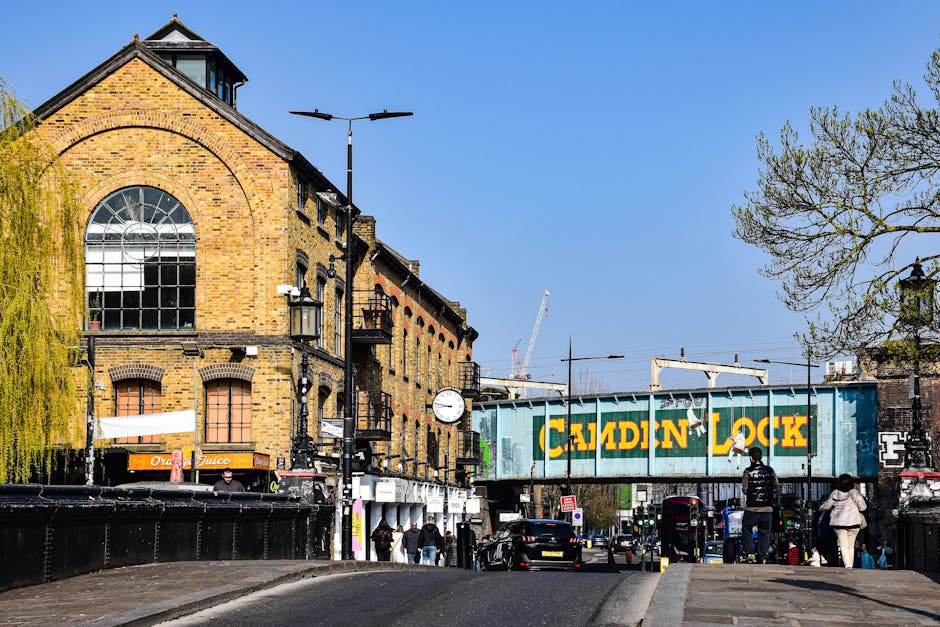 A busy street scene in Kentish Town during daytime featuring a large brick building with arched windows on the left, and a vintage railway bridge with a painted sign reading 'Camden Lock' spanning across the road. Several pedestrians are walking along the pavement, some carrying bags or waiting to cross. A black van is parked nearby, and a trolley loaded with cardboard boxes and wrapped furniture is seen on the pavement, indicating moving or packing activities. Streetlights, a clock, and trees, some with fresh green leaves, frame the scene against a clear blue sky. This image illustrates typical urban home relocation logistics, including loading and unloading furniture and packaging materials, as part of a professional removals service such as those offered by Removals Kentish Town, relevant to Camden Council rules for moving vans in Kentish Town.