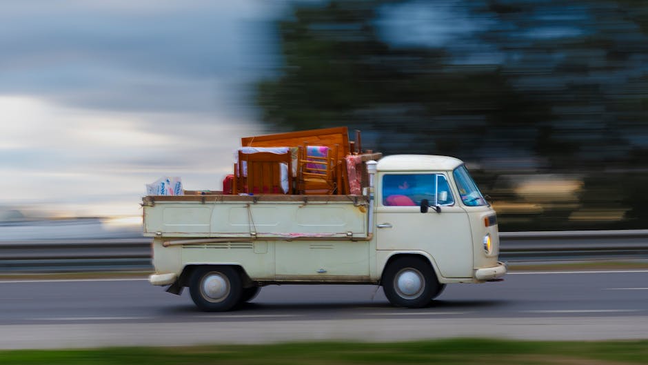 An older model cream-coloured van featuring a boxy design and small wheels is in motion along a highway, with a blurred background indicating high speed. The vehicle's flatbed cargo area is loaded with various household furniture items, including a wooden dining chair, a small wooden table, and a wooden cabinet, along with packed cardboard boxes and plastic bags. The furniture is secured with straps or padding, typical of home relocation or furniture transport. The scene suggests a moving process requiring careful loading and transportation, consistent with professional removals services such as those provided by Removals Kentish Town. The lighting is natural, and the background features motion-blurred trees and buildings, emphasizing the movement of the vehicle during a home relocation or moving service.