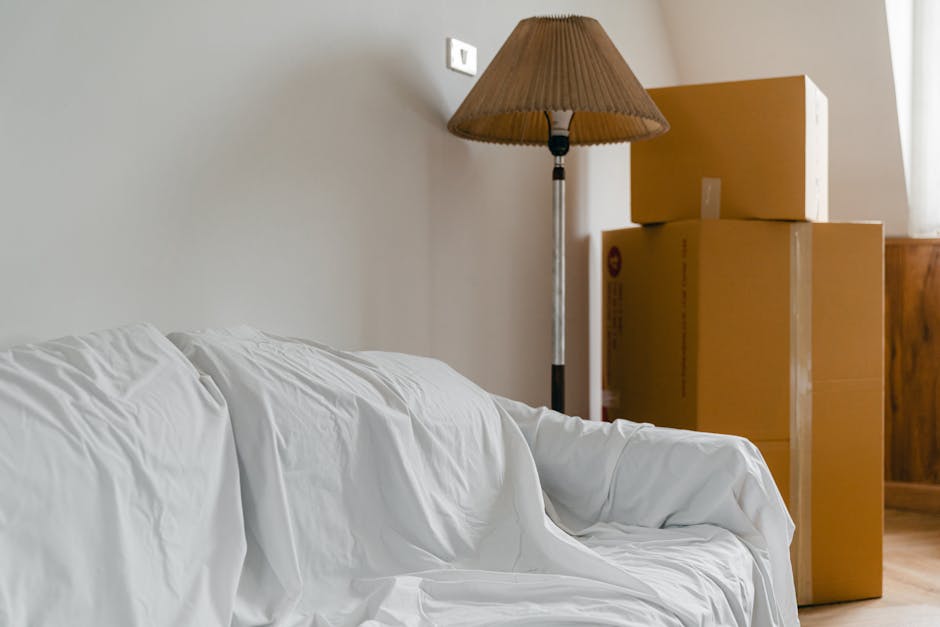 The image shows a living room corner with a white fabric-covered sofa positioned against a light grey wall. In the background, there are three cardboard moving boxes stacked near a window, indicating ongoing packing or a home relocation process. To the left of the boxes, a tall floor lamp with a beige pleated lampshade stands, casting soft light on the scene. The wooden flooring is visible beneath the sofa and boxes. The setting appears tidy, with the boxes likely prepared for transport, and no furniture or items are obstructing pathways, suggestive of an organized packing process often handled by services like Removals Kentish Town, which specialises in removals and moving logistics.