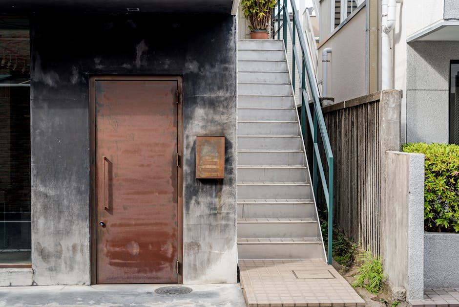 A close-up view of a narrow outdoor staircase leading up to a residential entrance, situated beside a weathered, dark grey concrete wall with a rusty metal door and a small, rectangular mailbox attached to it. The staircase is made of grey concrete with metal handrails painted turquoise, ascending from the pavement on the right side of the image. A potted plant is visible at the top of the stairs, and the small landing at the base of the staircase is paved with brick tiles. To the right, a section of a white building with a window and a concrete fence partially covered by greenery can be seen. The environment appears to be a narrow alleyway or side access pathway, typical of urban house locations, demonstrating the challenges of moving furniture and boxes through limited stair access. This image represents the typical context faced during house removals involving narrow staircases, where careful planning by removals Kentish Town may be necessary for efficient furniture transport.