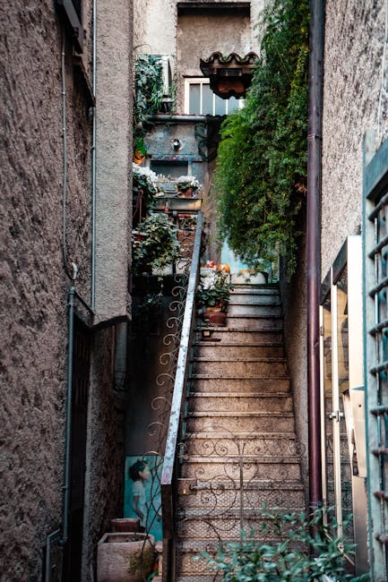 A narrow outdoor staircase made of concrete with visible wear and small decorative patterns on the risers, situated between two tall, textured exterior walls of buildings. The left wall is made of weathered stone or rough concrete, while the right wall appears stuccoed with partially visible blue painted sections. A black metal handrail with scrollwork detail runs alongside the stairs, supporting large flower pots filled with blooming plants and greenery placed on small ledges and along the steps. Potted plants and flowers in various containers, including terracotta and plastic, decorate the stairway, which leads upward beneath a small wooden roof or overhang. To the left at the bottom, a metal gate with vertical bars is partly closed, and in the background, the upper part of a window with a white frame and dark glass can be seen, along with additional greenery. The scene is illuminated by natural daylight, highlighting the textured surfaces and lush foliage, and depicts an authentic residential alleyway suitable for house removals or furniture transport in tight urban spaces.