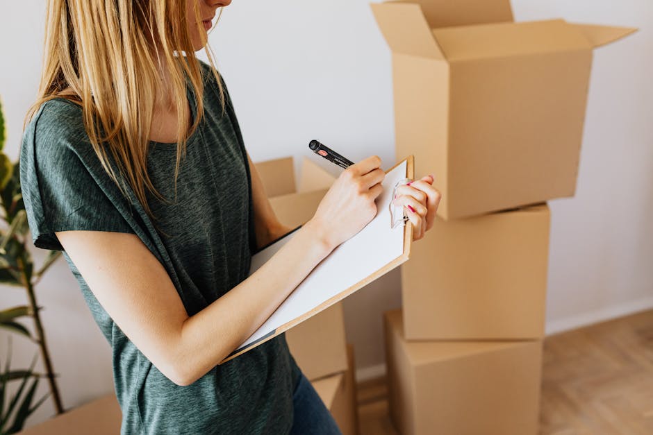 A woman with long blonde hair wearing a dark green T-shirt is inside a residence, standing near a stack of three cardboard moving boxes, with the largest box open at the top. She is holding a black marker and writing on a small notepad or clipboard, possibly creating a packing or inventory list as part of the home relocation process with the assistance of Removals Kentish Town. The room has a light wall and wooden flooring, with a potted plant partially visible on the left side of the image. The scene depicts an organized packing and moving stage, highlighting the preparation and planning involved in furniture transport and house removals, supported by professional removals services such as those offered by Removals Kentish Town.