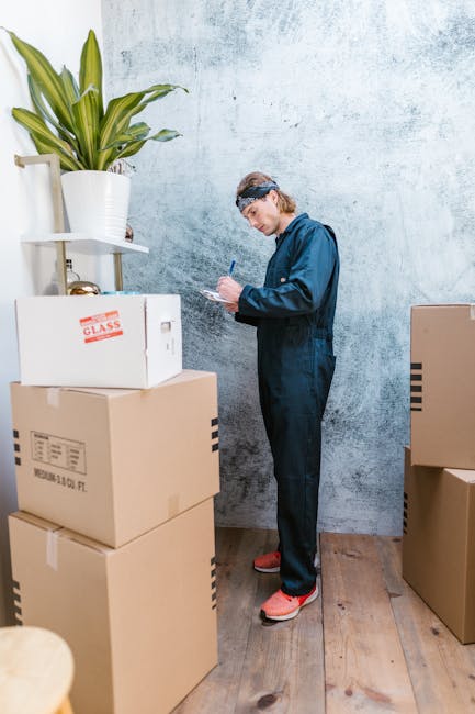 A man dressed in a blue jumpsuit and wearing a headband is standing indoors against a textured, light-colored wall. He is surrounded by several packed cardboard boxes, one of which has a label indicating fragile contents, and a large white plant pot with a green leafy plant on a shelf nearby. The boxes are stacked on a wooden floor, and some are positioned in front of him while others are to his side. The man is holding a pen and appears to be noting details on a clipboard or paper, part of a home relocation or packing process. The environment suggests an organized moving or packing stage within a residential space, with visible furniture and packaging materials. Removals Kentish Town offers professional services for house removals and furniture transport, supporting the logistics of packing and moving.