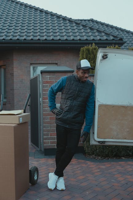 A man in casual clothing, including a baseball cap, gloves, a black padded vest, and blue sleeves, is standing next to an open vehicle door during a home relocation process. The vehicle appears to be a removal van parked on a paved driveway outside a residential property with a brick building and a tiled roof visible in the background. Several cardboard boxes are positioned nearby, indicating packing activities, and the man is observing or preparing to load or unload items such as furniture or boxes as part of the packing and moving process. This scene reflects professional furniture transport and home removal services offered by Removals Kentish Town, emphasizing efficient loading procedures in a residential environment.