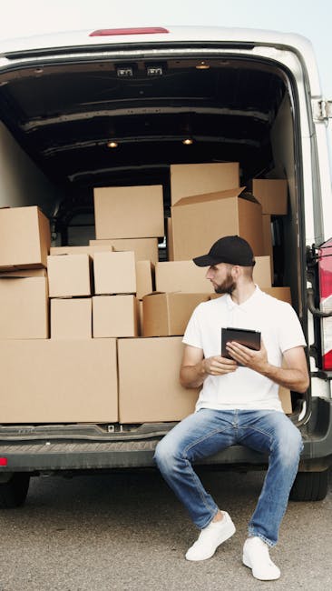 A man wearing a black cap, white t-shirt, and blue jeans is sitting on the pavement beside the open rear doors of a white van, which is loaded with multiple stacked cardboard boxes of various sizes. The boxes are sealed with packing tape and are arranged to maximize space within the vehicle, indicating a home relocation or furniture transport process. The interior of the van shows additional packing materials, possibly plastic wrappings or blankets, used to protect furniture and fragile items during moving. The man appears to be reviewing information on a tablet device, likely related to the loading process or moving schedule. The scene takes place outdoors, with the van positioned on a street or driveway, and the lighting suggests daytime conditions. This image is associated with removals services, such as those offered by Removals Kentish Town, highlighting the logistics involved in packing and loading household items for a move near NW5 or similar areas.