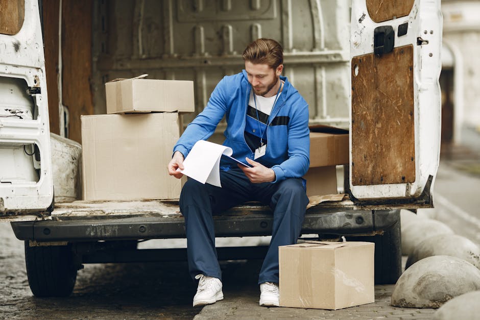 A man in casual clothing, including a baseball cap, gloves, a black padded vest, and blue sleeves, is standing next to an open vehicle door during a home relocation process. The vehicle appears to be a removal van parked on a paved driveway outside a residential property with a brick building and a tiled roof visible in the background. Several cardboard boxes are positioned nearby, indicating packing activities, and the man is observing or preparing to load or unload items such as furniture or boxes as part of the packing and moving process. This scene reflects professional furniture transport and home removal services offered by Removals Kentish Town, emphasizing efficient loading procedures in a residential environment.