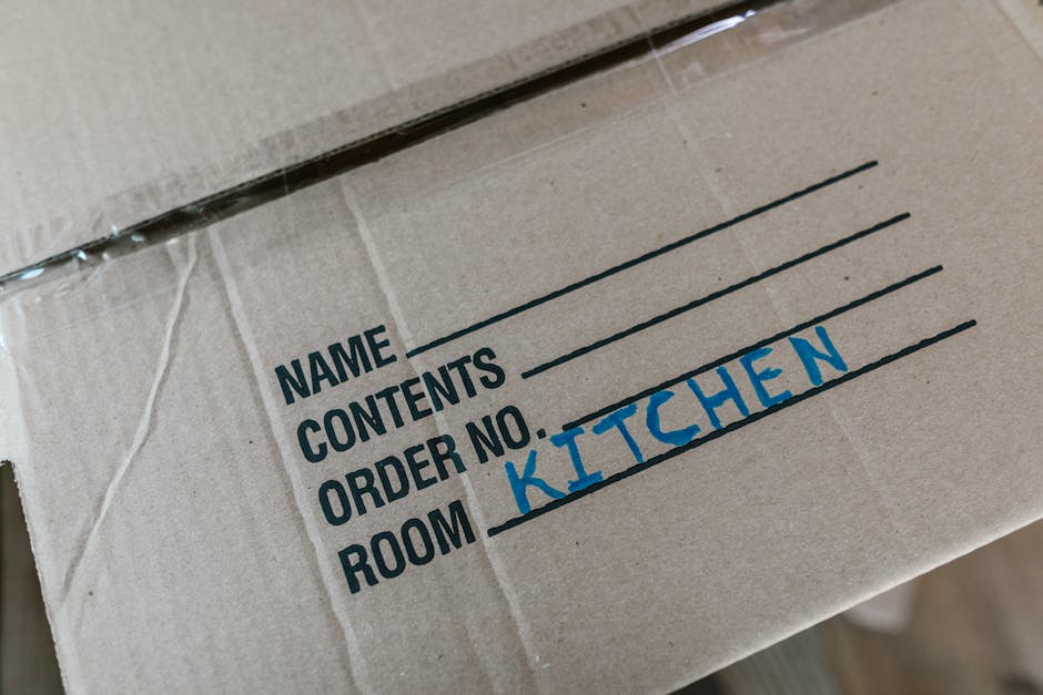 Close-up view of a cardboard moving box with printed labels indicating sections for home relocation. The label on the box shows fields for 'NAME,' 'CONTENTS,' 'ORDER NO.,' and 'ROOM,' with the word 'KITCHEN' handwritten in blue marker next to the 'ROOM' field. The box is wrapped in clear plastic tape along the edges to secure the contents. The background reveals additional packing materials, suggesting an organized packing process before furniture transport. The scene is set indoors, near an open doorway or loading area, with natural lighting highlighting the details of the packaging and preparing for the loading process, typical of house removals carried out by companies like Removals Kentish Town as part of their professional removals and moving services.