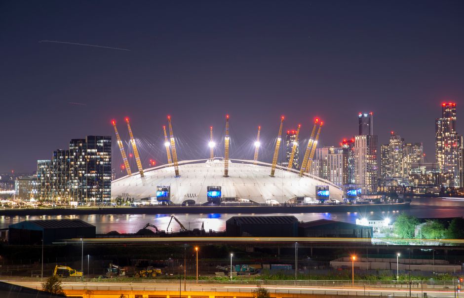 A nighttime cityscape featuring the O2 Arena in London, with its distinctive white, dome-shaped roof supported by large yellow cranes and numerous bright red warning lights on top of each crane. The arena is situated along the river Thames, whose calm waters reflect the illuminated building and city skyline in the background. To the right, a cluster of modern high-rise buildings with lit windows contribute to the city lights. In the foreground, construction equipment and vehicles are visible on the road and construction site, indicating ongoing development activities. The scene captures the vibrant urban environment, including lighting that highlights the structure and surroundings, relevant to house removals and moving logistics associated with projects like the O2 Forum move day plan, as coordinated by Removals Kentish Town for event-related relocations.