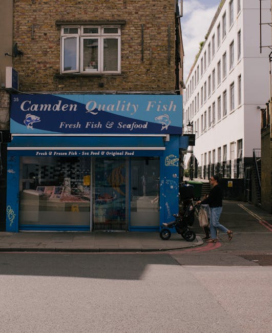 The image depicts the storefront of Camden Quality Fish, a seafood shop situated on a street corner in an urban area. The shop has a blue sign with white lettering that reads 'Camden Quality Fish' and 'Fresh Fish & Seafood,' and features a graphic of a fish. Large glass display windows showcase various seafood products inside, with some packaged on ice and others in containers. To the right of the shop, a person dressed in casual clothing is seen pushing a stroller along the pavement, near a woman carrying a bag. The surrounding environment includes residential and commercial buildings, with a brick building above the shop and a modern white building adjacent. The scene is lit by daylight, and the pavement appears clean and well-maintained. This image is relevant to house removals and moving services, highlighting the urban setting and typical street environment involved in relocation logistics, with [COMPANY_NAME] removing household items in the context of home relocation or furniture transport.