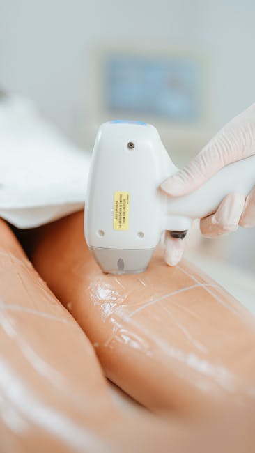 A close-up view of a professional moving technician from Removals Kentish Town using a handheld ultrasound device to scan a client's leg during a home relocation assessment. The technician wears a white glove and holds the device against the client's thigh, which is covered in a transparent gel to facilitate the scan. In the background, a blurred computer monitor displays a blue-toned interface, indicating a controlled environment for the evaluation process. The setting appears to be an indoor space with neutral tones, highlighting the professional approach to packing, moving, and health and safety procedures involved in house removals services in Kentish Town. The image emphasizes careful handling and diagnostic assessment as part of comprehensive moving logistics and packing strategies.