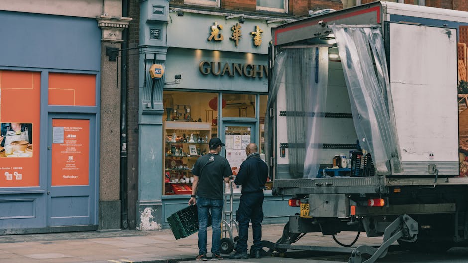 Two men are standing outside the storefront of Guang, a shop with traditional Chinese signage in gold Chinese characters and the word 'GUANG' displayed beneath. They are preparing for furniture transport or a home relocation, with one man holding a small wheeled dolly, and the other carrying a black plastic crate. A large moving truck with an open back is parked nearby on the pavement, partially covered with transparent plastic sheeting to protect the interior from the weather. The truck is positioned adjacent to the shop, ready for loading or unloading furniture or packaged items. The scene is set during daytime in an urban area with a mix of brick and plaster building facades, and the lighting is natural, illuminating the scene evenly. Removals Kentish Town is involved in house removals, evidenced by the presence of packing materials, boxes, and equipment suitable for furniture transport and professional moving logistics.