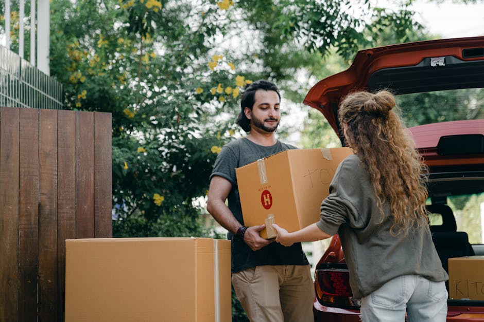 A man with shoulder-length dark hair and a beard is engaged in a home relocation process, standing outdoors beside a red vehicle with its trunk open. He is holding a large cardboard box sealed with pa